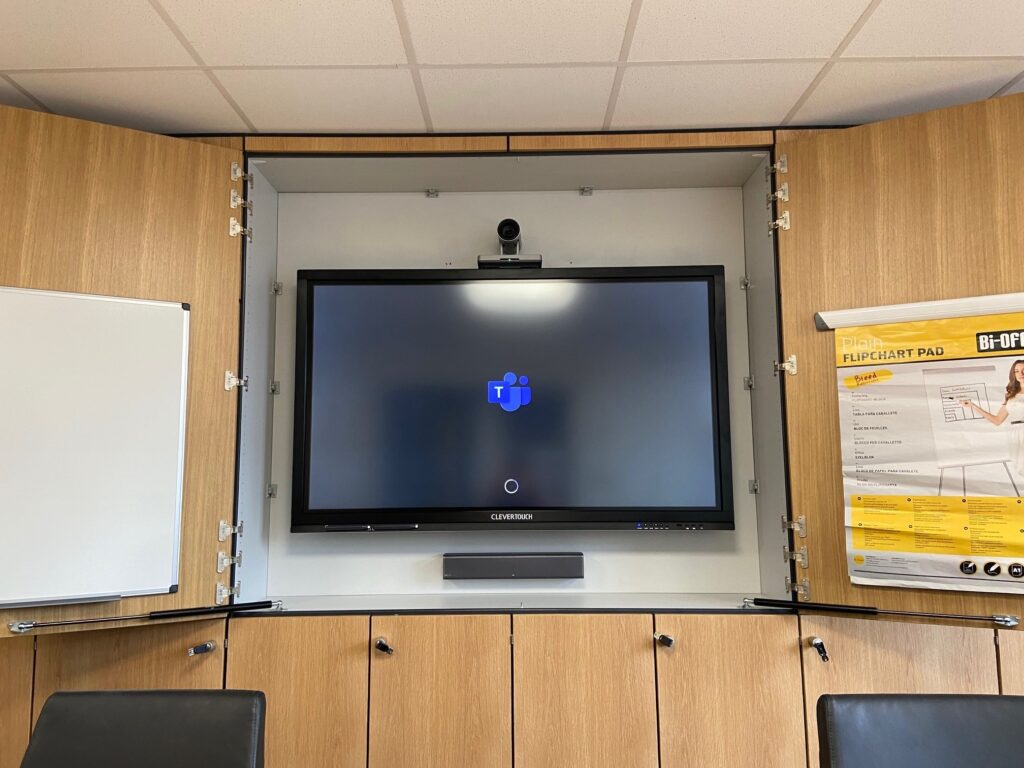 A conference room with audio visual systems including a large screen displaying a loading icon, flanked by whiteboards and mounted cabinets.