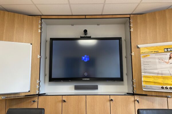 A conference room with audio visual systems including a large screen displaying a loading icon, flanked by whiteboards and mounted cabinets.