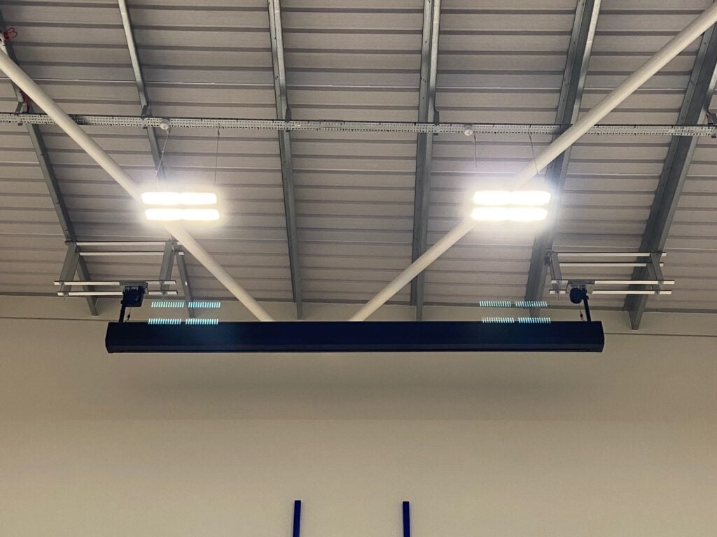 Ceiling view showing an industrial heater with bright lights and audio visual systems attached, mounted on gray beam structures in a warehouse.