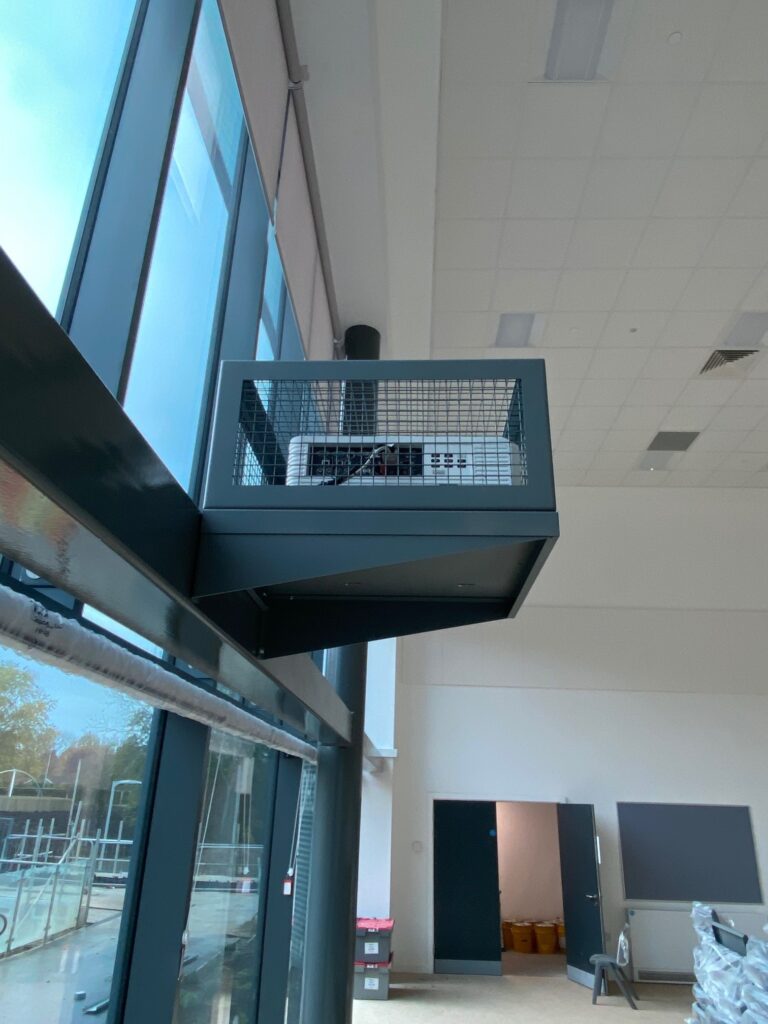 Modern industrial-style interior featuring a high ceiling, large windows, and a small elevated balcony with a metal railing, supported by a black structural beam.