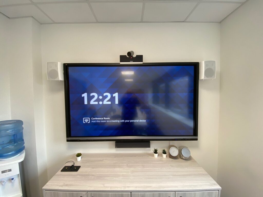 A modern conference room setup displaying a large screen showing the time "12:21" and a message about connecting to a meeting, flanked by two speakers, in a clean and organized space with advanced audio visual systems.