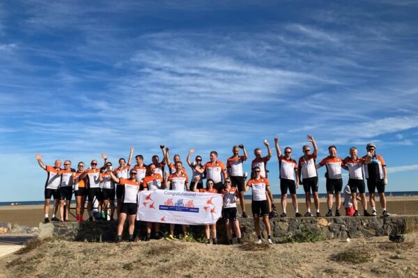 A group of cyclists celebrating on a beach, holding a congratulations banner with clear blue skies in the background.