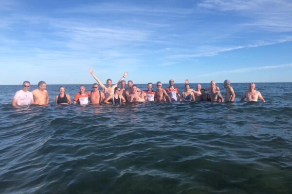 A group of people smiling and posing in the ocean, some waving, under a clear blue sky.