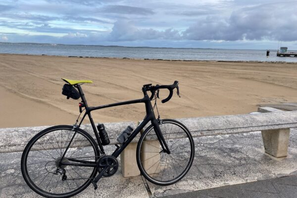 A black road bicycle leaning against a seaside railing with a sandy beach and cloudy sky in the background.