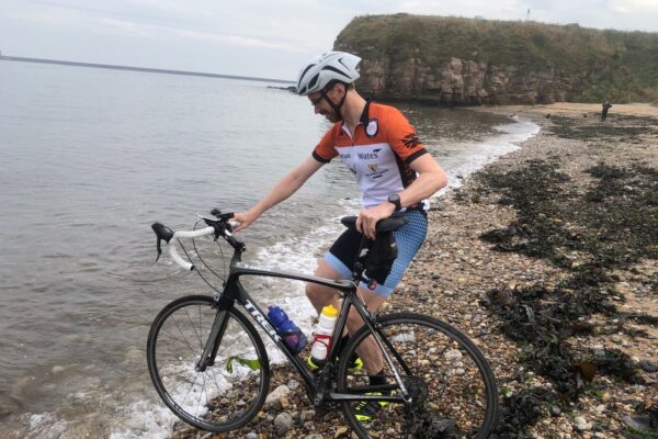 A cyclist in a helmet and sportswear stands next to a road bike on a pebbly beach, with cliffs and the sea in the background.