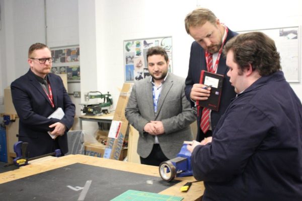 Four men in a workshop, one demonstrating a device to three others who are attentively observing.