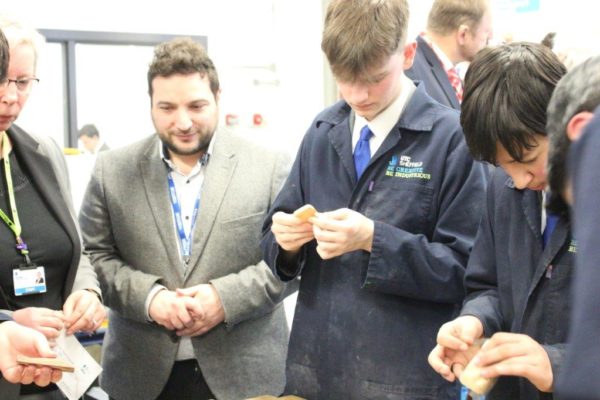 Two students and two adults examine a project in a classroom setting, with one student actively working on a small component.