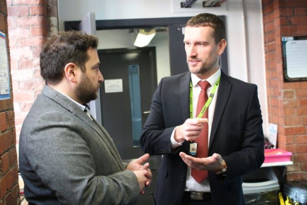 Two men in business attire having a conversation in an office hallway. one holds his id badge while gesturing with his hand.