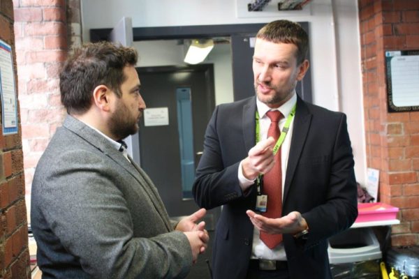 Two men engaged in conversation in a hallway, one holding a pen and wearing a badge, both dressed in business attire.